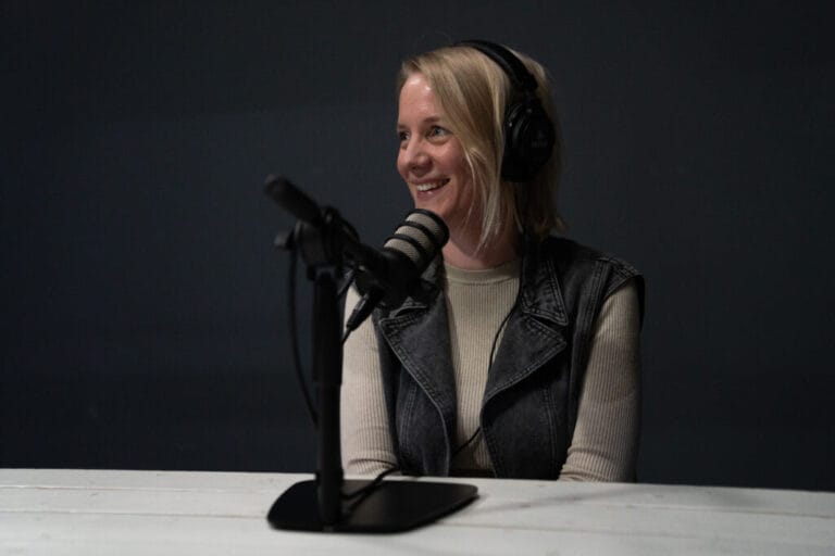 Vrouw met blond haar en koptelefoon op zit achter een podcastmicrofoon