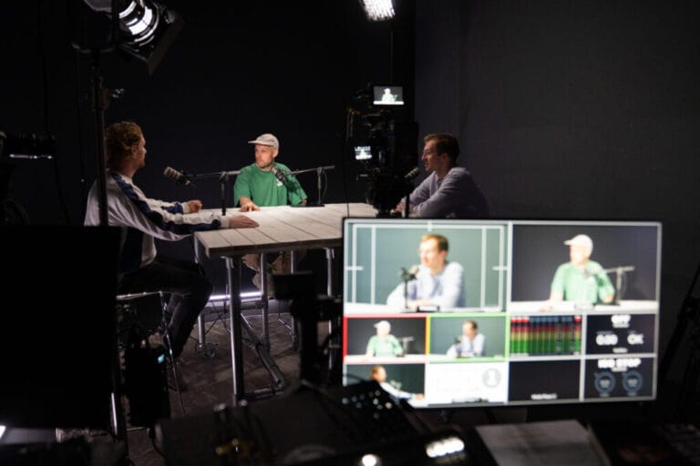 Drie mensen om een tafel nemen een podcast op. Op de voorgrond zie je een computerscherm met een videobeeld van elke spreker.